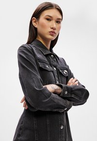 Jaqueta de ganga preta com colarinho, bolsos frontais e acabamentos prateados. Mangas longas, corte descontraído e detalhe de costura visível.