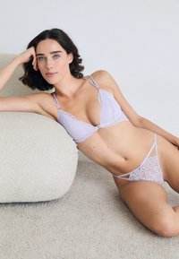 Vrouw die leunt op een gestructureerde crèmekleurige bank, gekleed in een lavendelkleurige kanten beha en bijpassende slip, tegen een effen lichtgrijze achtergrond.
