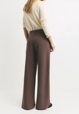 Kobieta stojąca tyłem, ubrana w kremowy sweter i brązowe spodnie z wysokim stanem i szerokimi nogawkami oraz czarne buty na białym tle.