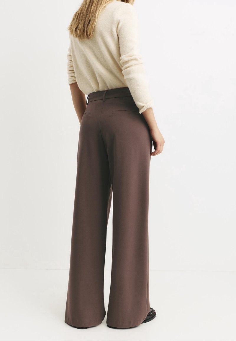 Femme debout, dos à la caméra, portant un pull crème et un pantalon marron taille haute à jambes larges avec des chaussures noires sur fond blanc.