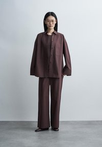 Femme aux longs cheveux noirs et lunettes portant une chemise marron ample à boutons, un pantalon large assorti et des chaussures marron, debout sur un sol gris.