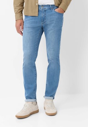 Mand iført lyseblå opkørte jeans, beige jakke, hvide sneakers, stående med en hånd i lommen mod hvid baggrund.