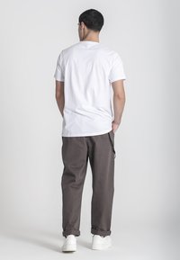Maglietta bianca in cotone a maniche corte, abbinata a pantaloni marroni larghi e scarpe da ginnastica bianche, vista da dietro.