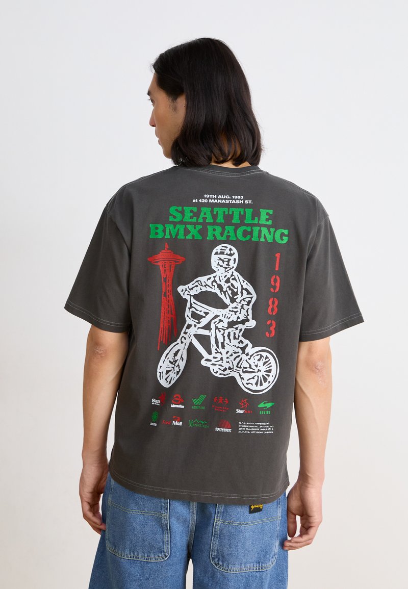 T-shirt oversize gris avec un imprimé graphique d'un rider BMX, de la Space Needle de Seattle, et du texte en vert et rouge, avec divers logos de sponsors en dessous.