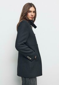 Cappotto di lana grigio scuro con silhouette strutturata, colletto alto e bottoni decorativi sui polsini. Due tasche laterali visibili.