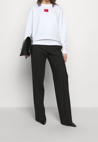 Sudadera blanca con logo rojo, combinada con pantalones amplios negros y tacones negros de punta. Modelo sosteniendo un bolso clutch negro.