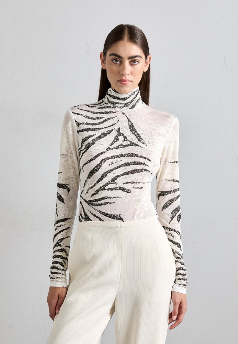 Duga rukava, majica visokog vrata od prozirnog materijala s crnim zebrinim prugama na svijetloj pozadini, u kombinaciji s kremasto bijelim plisiranim hlačama.