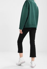 Persoon in een donkergroene oversized sweatshirt, zwarte cropped broeken en witte high-top sneakers, staand tegen een effen achtergrond.