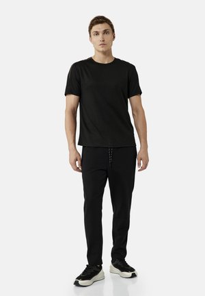 Giovane uomo in piedi che indossa una maglietta nera semplice a maniche corte, pantaloni neri con coulisse e scarpe da ginnastica nere e bianche su sfondo bianco.