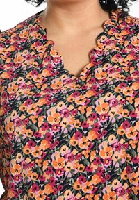 Donna che indossa una blusa floreale con fiori arancioni, rosa e viola e un colletto scallop su uno sfondo bianco.