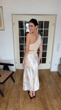 Kobieta uśmiechająca się przez ramię, ubrana w beżowy top na jedno ramię i białą satynową spódnicę w kwiaty, stojąca na drewnianej podłodze przed drzwiami z przeszklonymi panelami.
