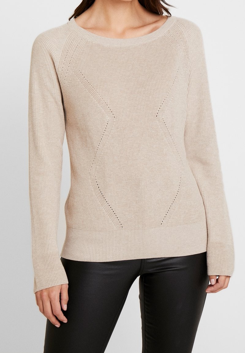 Kobieta w beżowym swetrze dzianinowym z subtelnymi wzorami i długimi rękawami, w zestawieniu z czarnymi skórzanymi spodniami, widoczna od szyi do bioder.