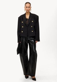 Blazer noir croisé à double boutonnage avec des boutons dorés, associé à un pantalon large en similicuir noir brillant. Accentué par une pochette noire texturée et des talons.