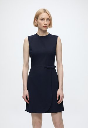 Femme aux cheveux blonds courts portant une robe bleu marine foncé sans manches, longueur au genou, avec un petit détail de ceinture sur un fond blanc uni.