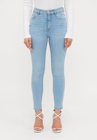 Ljusblå högmidjade skinny jeans med ett subtilt urtvättat mönster, femficksdesign och detaljerad bakgördel. Vita sandaletter med remmar är synliga.