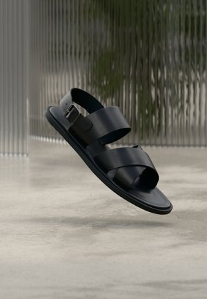 Schwarze Ledersandale mit zwei breiten Riemen und einer Schnalle, die über einem polierten Betonboden vor einem unscharfen, geriffelten Glas-Hintergrund schwebt.