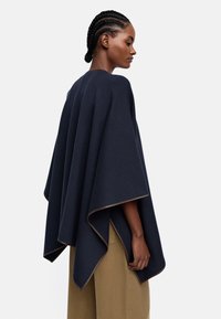 Granatowy wełniany ponczo o luźnym kroju, z gładką teksturą i kontrastowym beżowym wykończeniem na krawędziach. Bocznie rozcięcia dla swobody ruchów.