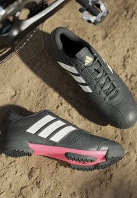 Ein Paar schwarze Fahrradschuhe mit weißen Streifen und pinken Sohlen, die auf Erde neben einem Fahrradpedal und Kettenblatt platziert sind.