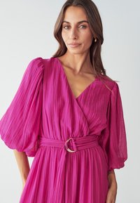 Fuchsia Wickelkleid aus strukturiertem Stoff, mit puffigen Ärmeln und tailliertem Schnitt, ausgestattet mit einer runden Gürtelschnalle.