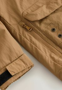 Giacca leggera marrone con zip frontale, chiusure a scatto e dettagli in velcro sui polsini, con una finitura in tessuto a trama.