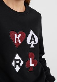 Svart sweatshirt med flerfärgade, strukturerade applikationsdesigns av spelkortssymboler: hjärta, spade, diamant och klubba med bokstäver.