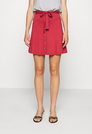 Czerwona spódnica w kształcie litery A z paskiem do wiązania, zapinana na guziki z przodu, o gładkiej fakturze i długości do kolan. Dobraną do niej są klapki w panterkę.