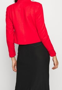 Kvinna iklädd en ljus röd kort jacka och en svart kjol med hög midja, sedd bakifrån mot en enfärgad bakgrund.