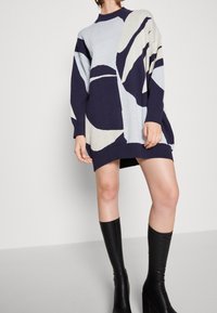 Abito in maglia con scollo rotondo e maniche lunghe, in blu navy e azzurro con forme astratte. Abbinato a stivali neri al ginocchio.