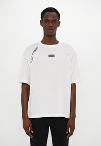 T-shirt blanc à coupe décontractée, manches courtes, découpes sur les côtés et patch logo sur la poitrine. Présente des accents texturés sur les épaules.