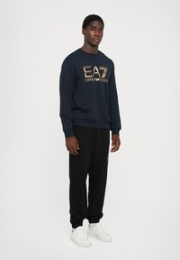 Navy sweatshirt with gold EA7 logo, paired with black joggers and white sneakers. The fabric is smooth with ribbed cuffs and hem.
