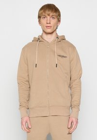 Beige zip-up hette i stoff med snorer, frontlommer, ribbede mansjetter og subtil merking på venstre bryst. Glatt tekstur.