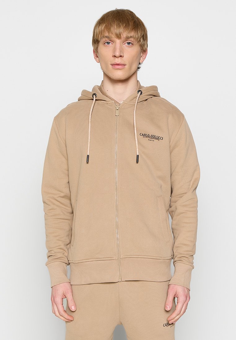 Beige zip-up hette i stoff med snorer, frontlommer, ribbede mansjetter og subtil merking på venstre bryst. Glatt tekstur.