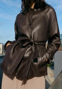 Brązowa skórzana kurtka z paskiem w talii, z zaokrąglonym kołnierzem, zapięciami na guziki i kontrastującymi przeszyciami. Gładka faktura z kieszeniami.