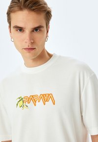 Hvid bomulds t-shirt med orange "PAPAYA" tekst og grøn papaya frugt grafik. Korte ærmer og klassisk crew neck design.