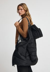 Svart puffjacka med kviltad design och flera fickor, bärs över en brun turtle neck. Har avtagbar huva och justerbara remmar.