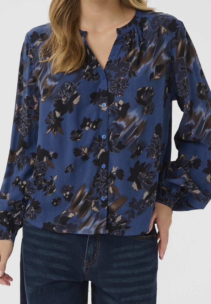 Femme portant un chemisier bleu à fleurs à manches longues avec des boutons sur le devant et un jean bleu foncé sur un fond uni.