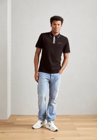 Polo negro de manga corta con ribete blanco, que cuenta con un cuello abotonado y un logo. Combinado con unos jeans de mezclilla azul claro y zapatillas blancas.