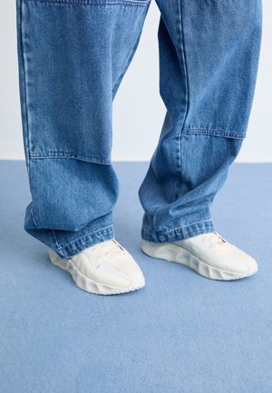 Witte sportieve schoenen met gestructureerde rubberen zolen, gecombineerd met losvallende blauwe denim broek. Achtergrond is een zachte blauwe ondergrond.