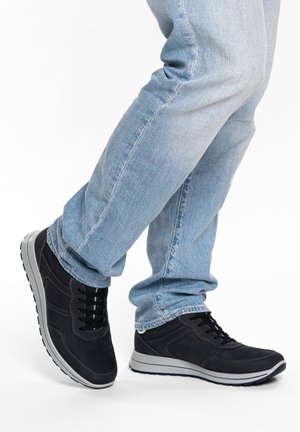 Persoon in lichtblauwe spijkerbroek zet een stap vooruit, met zwarte veter-sneakers met witte zolen op een witte achtergrond.