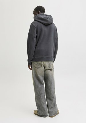 Man wearing a dark gray hoodie and loose faded gray jeans, standing facing away on a plain light background.