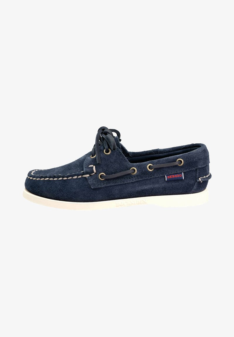 Bateau en daim bleu marine avec semelle blanche, détails cousus et œillets en laiton. Présente une fermeture à lacets et une petite étiquette rouge sur le côté.