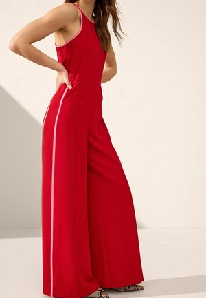 Vrouw in een mouwloze rode jumpsuit met wijde pijpen en roze zijstrepen, met schoenen met slangenprint, staand tegen een lichte achtergrond.