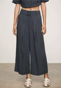 Pantalones de pierna ancha en tela gris oscuro con cintura ceñida y pliegues. Combinados con sandalias blancas de tiras. Textura suave.