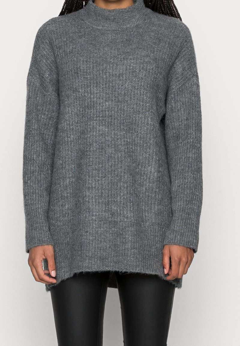 Personne portant un long pull gris en maille côtelée avec un col montant et un pantalon en cuir noir, debout devant un fond uni.