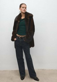 Brown fur coat over a green top, paired with dark denim jeans and black ankle boots. Features a wide collar and a gold belt buckle.