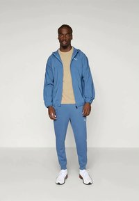 Man standing wearing blue Puma tracksuit, beige t-shirt, and white sneakers against plain light background.