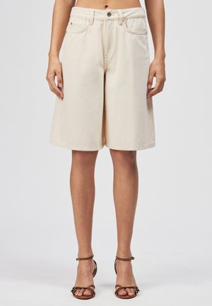 Osoba nosząca kremowe szorty dżinsowe do kolan z wysokim stanem oraz brązowe sandały na wysokim obcasie z paskami, stojąca na tle szarego tła.