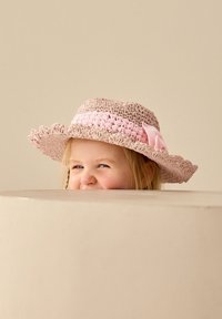 Chapeau de paille à larges bords ondulés en rose clair et beige avec un nœud rose, partiellement visible au-dessus d'une surface de couleur neutre.