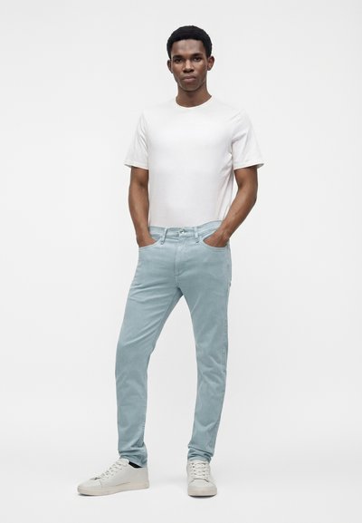 Jeune homme debout les mains dans les poches, portant une chemise blanche à manches courtes, un jean bleu clair et des baskets blanches sur un fond uni.