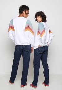 Hvid sweatshirt med flerfarvede diagonale striber i blå, pink og gul, parret med mørkeblå jeans og røde sko.